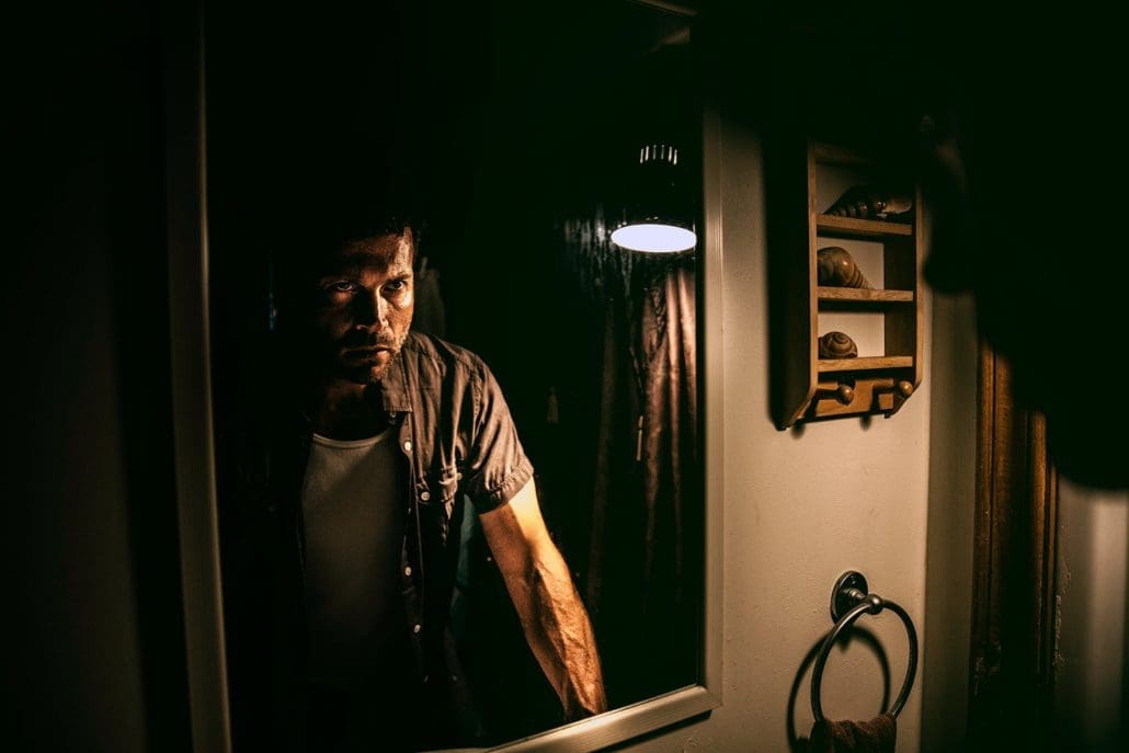 A man stands in a dimly lit bathroom, staring intensely at his reflection in the mirror. Shadows cover part of his face, creating a dramatic and moody atmosphere—a moment that hints at seeking support like Melbourne Counselling Services.