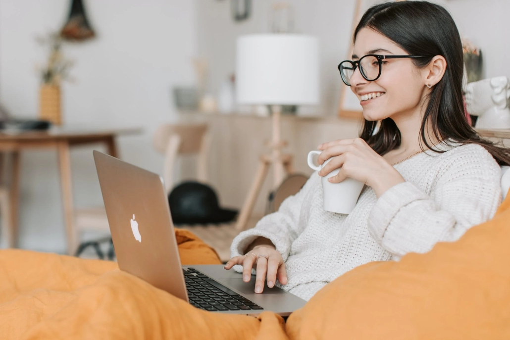 A woman wearing glasses and a cozy sweater smiles while holding a white mug and using her laptop, sitting comfortably with a yellow blanket in her bright, modern living room after connecting with Melbourne Counselling Services online.