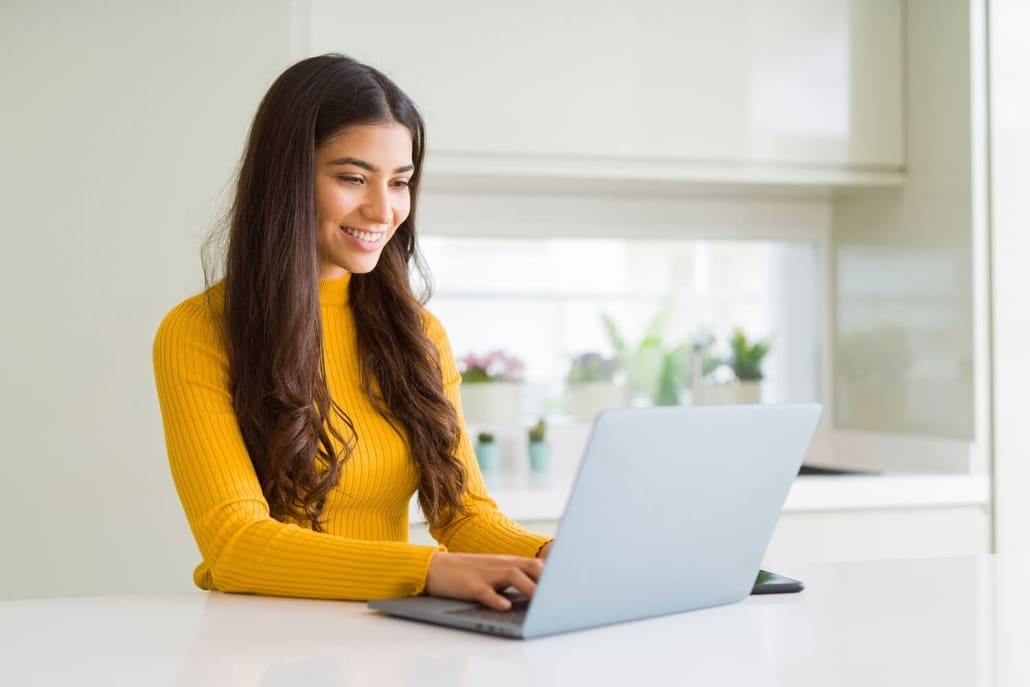Online Counselling Services A woman with long brown hair, wearing a yellow sweater, smiles while working on a laptop at a white table in a bright, modern kitchen. She searches for Melbourne Counselling Services as several potted plants decorate the background.